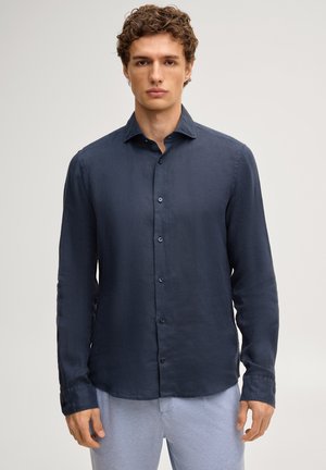 Jonge man draagt een donkerblauw, langärmelig overhemd met knopen en een lichtgrijze broek, staand tegen een eenvoudige lichte achtergrond.