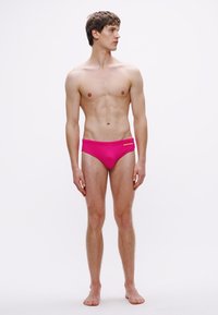 Ροζ μαγιό βερμούδα με απαλό ύφασμα και ελαστική ζώνη μέσης, με διακριτικό κίτρινο λογότυπο στο μπροστινό μέρος. Το μοντέλο στέκεται προφίλ.