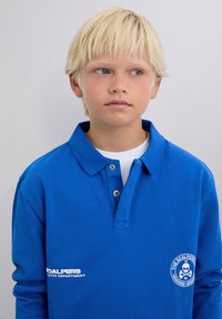 Niebieska koszulka polo z bawełny, z kołnierzykiem, trzema guzikami oraz białym haftowanym logo na piersi. Gładka tekstura, luźny krój.