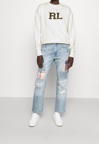 Bílá mikina s logem "RL" v hnědé barvě. Světle modré džíny s opotřebovaným vzhledem a nášivkami v kostkovaném a jednobarevném provedení. Noste je s bílými teniskami.