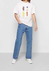 Vitt bomullströja med färgglada brandpostgrafiker; kombinerad med ljusblå denimjeans och vita sneakers med gula och blå detaljer.