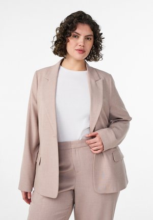 Kobieta o kręconych włosach ubrana w beżowy garnitur i biały top, stojąca na tle jednolitego białego tła.