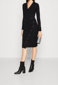 Donna in un vestito a wrap nero a coste di lunghezza media con bottoni laterali, stivali ankle neri con tacco, che tiene una pochette tessurizzata argentata.