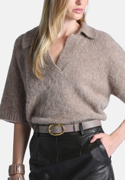 Femme portant un pull beige duveteux à manches courtes, rentré dans un pantalon en cuir noir avec une ceinture taupe et une boucle ovale dorée.