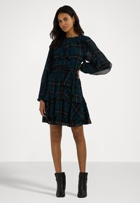 Grønn og marineblå rutete kjole med lange ermer, volangskjørt og knytting i halsen. Brukt med svarte ankelstøvler. Glatt stofftekstur.