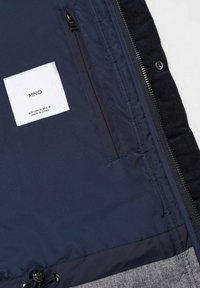 Granatowa kurtka zewnętrzna o gładkiej fakturze, zapinana na zamek, z wewnętrzną kieszenią i widoczną metką pielęgnacyjną. Szary detal z tkaniny u dołu.