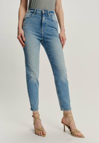 Mujer con jeans de mezclilla azul claro de cintura alta y sandalias de tacón alto de tiras beige, de pie contra un fondo liso.