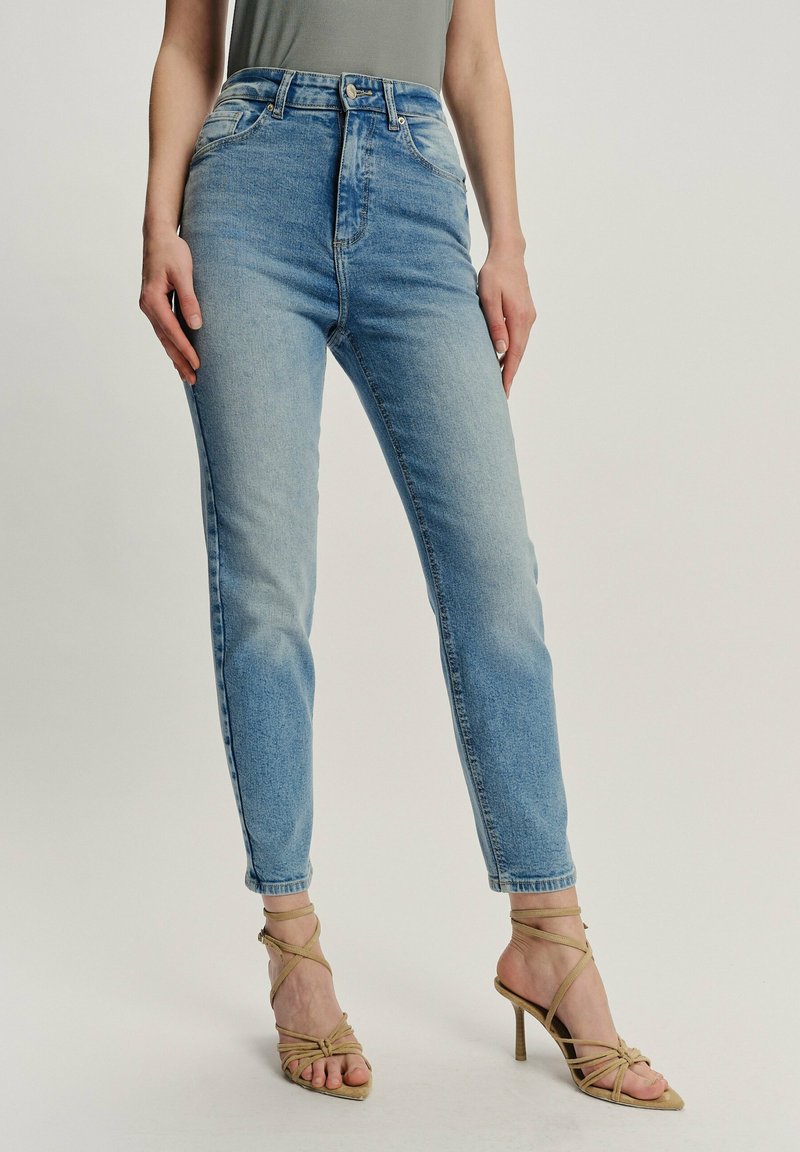 Mujer con jeans de mezclilla azul claro de cintura alta y sandalias de tacón alto de tiras beige, de pie contra un fondo liso.