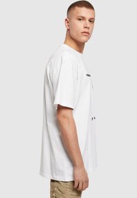 Hvid bomulds T-shirt med korte ærmer, rund hals, løs pasform, med sort tekst trykt på fronten. Glat tekstur og minimalistisk design.