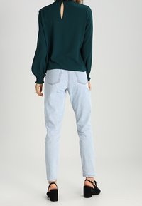 Blusa verde smeraldo a maniche lunghe con schiena a chiave, abbinata a jeans a vita alta di colore blu chiaro e scarpe con tacco blocco nere.