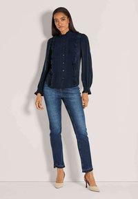 Marinebluse mit Rüschendetails, hohem Kragen und Knopfschluss; kombiniert mit dunklen Jeans und beigen spitzen Pumps. Glattes Material.