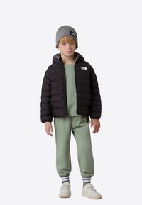 Čierna prešívaná bunda s kapucňou, zelená mikina, zladené nohavice a sivá pletená čiapka; so znakmi bieleho loga a textúrovanou tkaninou.