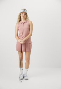 Paita sansi polo dengan lengan tanpa, rok lipit berwarna merah muda muda, dipadukan dengan kaus kaki pergelangan kaki putih dan sepatu atletik, sambil memegang klub golf.
