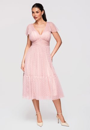 Kvinde iført en lyserød, prikprikket midi-kjole med flutterærmer og cremefarvede stiletter med ankelrem, står med den ene hånd på hoften.