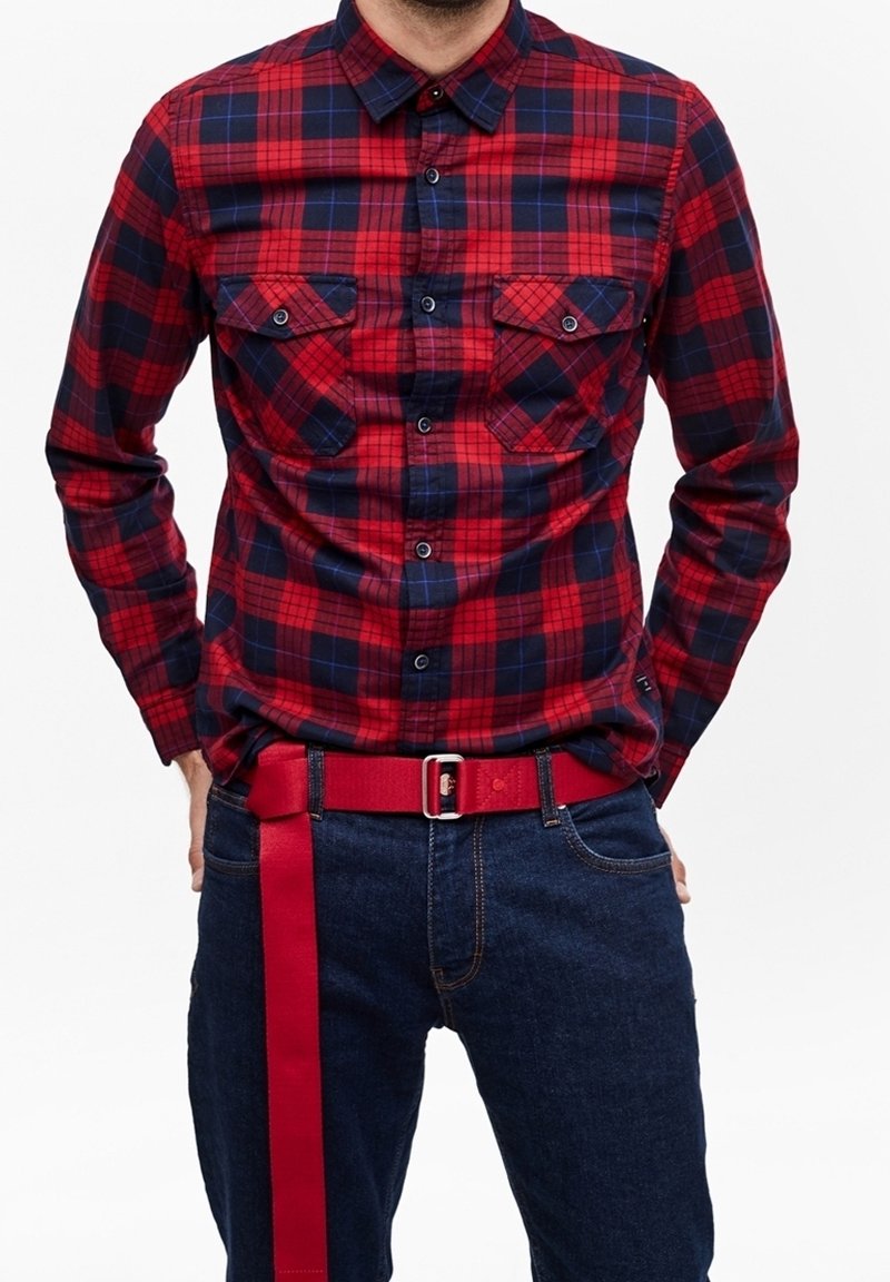 Homme portant une chemise à carreaux rouge et bleu marine avec deux poches poitrine et un jean bleu foncé avec une ceinture en tissu rouge pendante.
