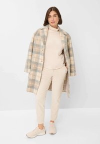 Manteau plaid beige avec une texture en laine naturelle, superposé à un col roulé beige clair et un pantalon en velours côtelé crème, accompagné de baskets beiges.