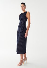 Marineblauwe mouwloze jurk met verticale witte strepen, taillering en strikdetail. Gecombineerd met witte gespenhoge haksandalen.