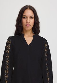 Blusa nera con scollatura a V e dettagli in pizzo intricati sulle maniche. Tessuto morbido con una vestibilità rilassata e una sottile texture.