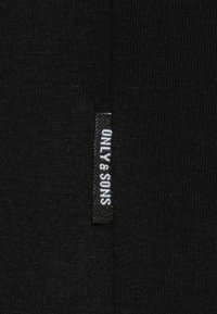Schwarzes Stoffetikett mit strukturierter Oberfläche und weißem Text "ONLY & SONS", der vertikal auf der linken Seite angebracht ist.