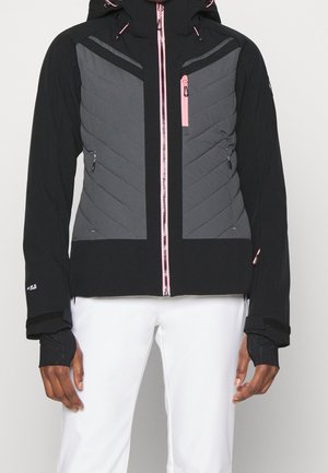 Schwarze und graue Jacke mit Reißverschluss und pinken Reißverschlussakzenten, Chevron-Steppung auf der Vorderseite, Daumenlöcher an den Ärmeln, getragen mit weißen Hosen.