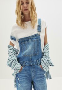 Blonde vrouw in een witte T-shirt en gestreept shirt dat van de schouders is afgezakt, gekleed in een blauwe spijkeroverall met haar handen in de zakken.