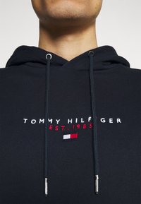 Sudadera navy con cordones, que presenta el texto bordado en blanco y rojo "TOMMY HILFIGER EST. 1985" y un pequeño parche del logo. Textura suave.
