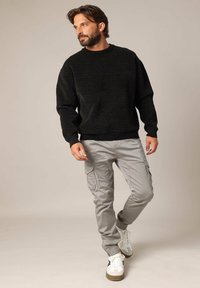 Homme avec une barbe portant un pull noir, un pantalon cargo gris et des baskets blanches, se tenant contre un fond beige uni.