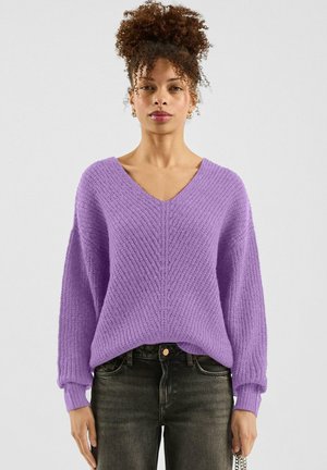 Fioletowy sweter na drutach z dekoltem w serek, szerokimi rękawami i teksturowanym wzorem. Połączony z ciemnymi jeansami, prezentującymi luźny krój i swobodny styl.