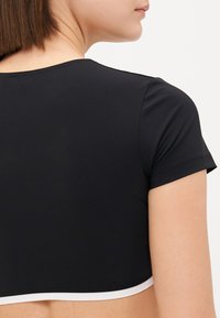 Fekete, rövid ujjú, crop top sima textúrával, lekerekített dekoltázzsal és kontrasztos fehér díszítéssel az alsó szegélyen. Hátulnézet látható.