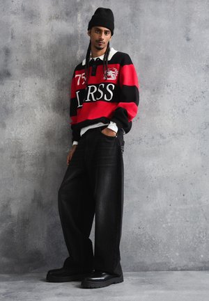 Mladý muž s vrkočmi, oblečený v čiernej čiapke, červeno-čiernom pruhovanom svetri, širokých čiernych džínsoch a čiernych topánkach, stojaci pri šedej stene.