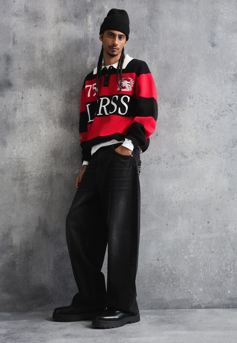 Junger Mann mit Zöpfen, der eine schwarze Mütze, einen rot-schwarz gestreiften Pullover, weite schwarze Jeans und schwarze Stiefel trägt und vor einer grauen Wand steht.