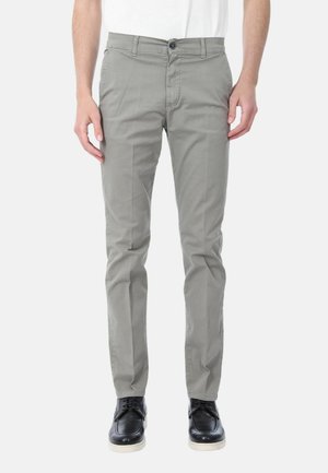 Uomo con pantaloni slim fit grigio chiaro e scarpe di pelle nere, in piedi davanti a uno sfondo bianco uniforme.
