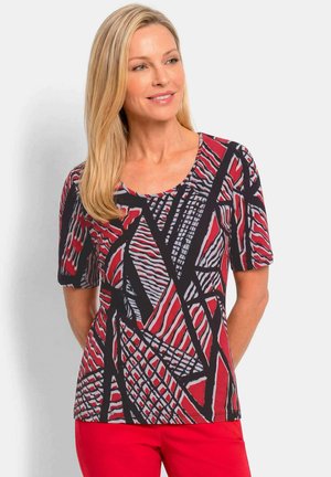 Blonde vrouw draagt een rood, zwart en grijs geometrisch patroon kort mouwtopje met rode broek, glimlachend en licht naar rechts kijkend.