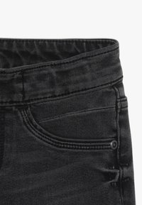 Jeans en denim noir avec une taille haute, un design à cinq poches, un léger effet déchiré et des détails en métal au niveau de la taille.