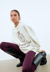 Felpa bianca con testo del logo marrone, abbinata a leggings bordeaux aderenti e stivali neri al ginocchio su uno sfondo neutro.