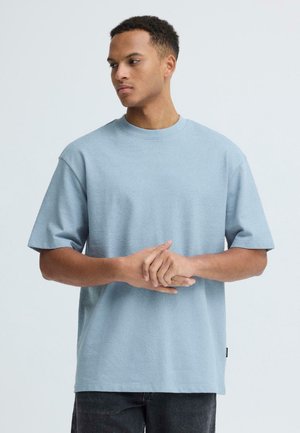 Jonge man met kort krullend haar, gekleed in een los lichtblauw shirt met korte mouwen, die zijn handen samenklapt en zijwaarts kijkt.