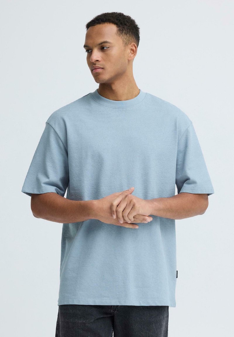 Νεαρός άνδρας με κοντά σγουρά μαλλιά που φοράει ένα φαρδύ, ανοιχτό γαλάζιο μπλουζάκι με κοντά μανίκια, ενώ σφίγγει τα χέρια του και κοιτάζει προς τα πλάγια.