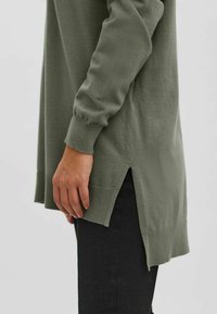 Langärmeliger, olivgrüner Pullover aus weichem Material mit gerippten Bündchen und seitlichen Schlitzen. Getragen über dunkler, enger Hose.