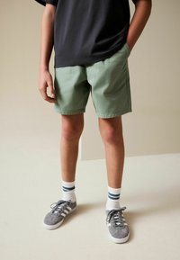 Pantaloncini di cotone verde chiaro con tasche laterali, abbinati a scarpe da ginnastica grigie e calzini bianchi con strisce blu, indossati da una persona dalla vita in giù.