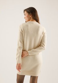 Beige sweater dress with long sleeves, featuring a rounded neckline and decorative beads on the shoulders. Ribbed hem and cuffs.