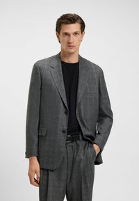 Blazer grigio a fantasia con chiusura a un solo bottone, due tasche frontali e una vestibilità rilassata, abbinato a una camicia nera e pantaloni coordinati.