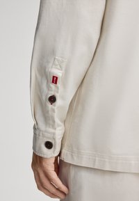 Crème-kleurig, textuurstof shirt met een geknoopte manchette en een rode accenttag met een uitroepteken.