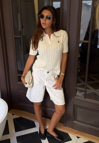 Femme portant des lunettes de soleil, haut blanc à manches courtes en maille torsadée, short blanc, chaussures noires, tenant une pochette beige, debout à l'intérieur près de portes vitrées.