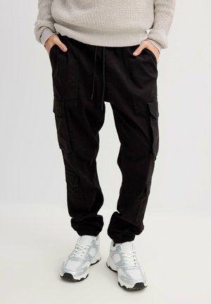 Pantalones cargo negros con puños fruncidos, que incluyen múltiples bolsillos laterales y una cintura elástica, combinados con zapatillas blancas y plateadas.