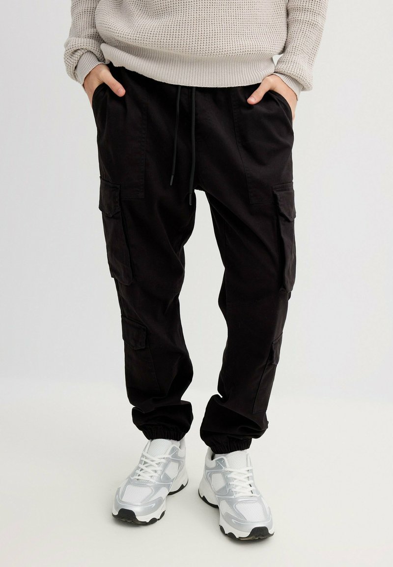 Pantalones cargo negros con puños fruncidos, que incluyen múltiples bolsillos laterales y una cintura elástica, combinados con zapatillas blancas y plateadas.