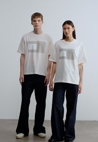 Λευκά βαμβακερά μπλουζάκια με γκρι γραφικό "TOMMY JEANS", συνδυασμένα με σκούρα μπλε τζιν παντελόνια. Τα μοντέλα στέκονται μπροστά σε ουδέτερο φόντο.