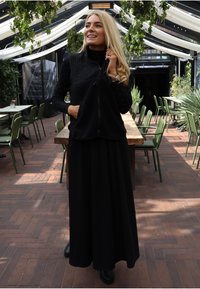 Fekete ujjatlan polár mellény elején cipzárral, fekete hosszú szoknya felett. A mellény texturált anyagból készült, finom fényes díszítésekkel.