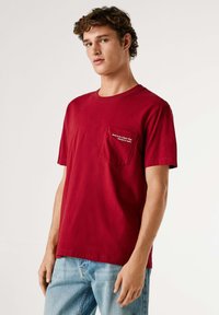 Rood katoenen T-shirt met een ronde hals en korte mouwen, voorzien van een borstzakje aan de linkerkant met geborduurde tekst. Gecombineerd met lichtblauwe spijkerbroek.