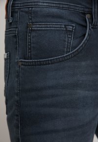 Los pantalones vaqueros de mezclilla azul oscuro cuentan con cinco bolsillos, costuras sutiles y acentos de metal. El material tiene una textura suave con un ligero brillo.