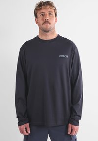 T-shirt noir en coton à manches longues avec col rond et logo bleu subtil sur la poitrine. Coupe ample avec ourlet droit et texture douce.